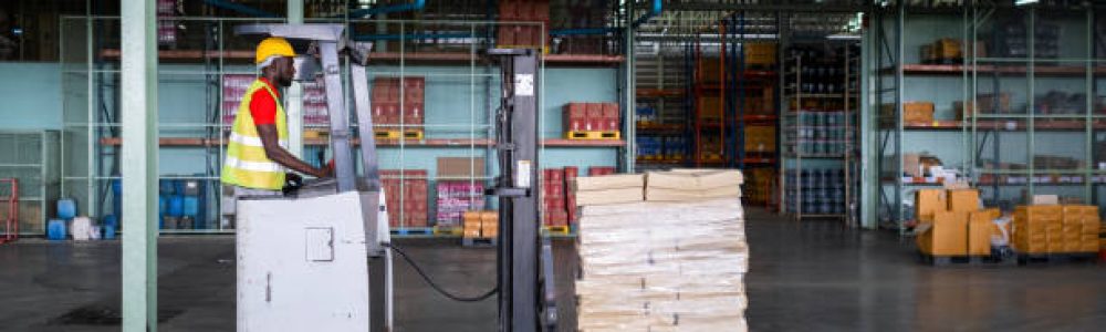 African American workers drive forklifts which holding many boxes with safety helmets and vests in the automotive parts warehouse. Transportation, delivery, and industrial factory concept.